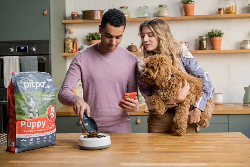 A young couple in their kitchen holding their dog while they use the PitPat app to work out how PitPat Food to feed their dog.
